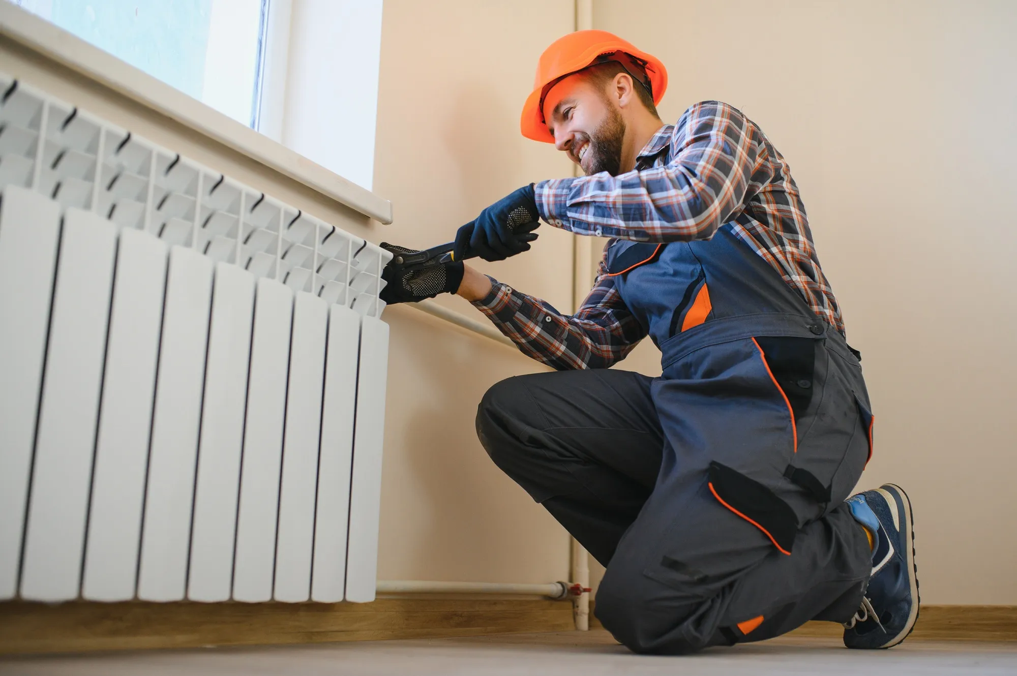Radiator repair in Cheverly, Maryland: Smiling worker in hard hat checking a white radiator. Plumber fixing heating system in home.
