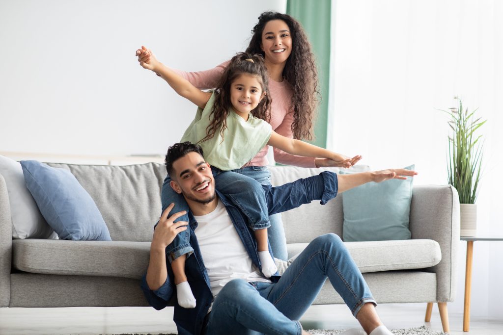Happy family enjoying time indoors, with a child on her father's shoulders, arms outstretched, mother standing behind. Emergency Heating in Cheverly, Maryland.