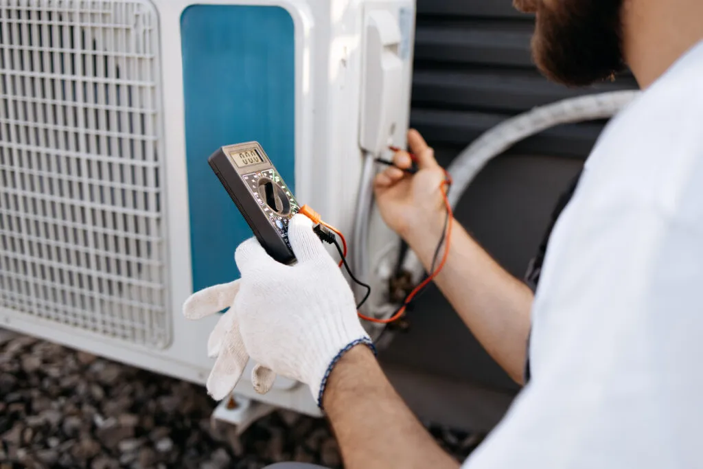 HVAC technician in Cheverly, Maryland, wearing gloves, tests an air conditioner unit with a multimeter, ensuring optimal performance for Hyattsville homes.
